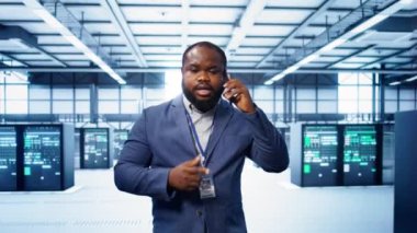 Bilgi işlem teknisyeni veri merkezinde telefonla konuşuyor, sunucu bakımını ve sorun çıkarma görevlerini koordine ediyor. Sunucu rafları ve yüksek teknolojili ekipmanlarla çevrili bir yönetici. Telefon görüşmesi, kamera B