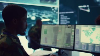 Ordu subayı, askeri komuta merkezinde gerçek zamanlı uydu istihbaratını inceleyerek, sahadaki beklenmedik gelişmeleri gidermek için acil durum planları geliştiriyor. Asker düşman sunucularını izliyor. Kamera B.