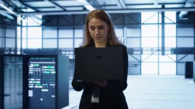 Veri merkezinde yürüyen yazılım geliştiricisi, laptop kullanarak sunucu işlemlerini denetliyor. Not defteri, kamera A kullanarak sunucu çiftliğinde ideal performans ve veri bütünlüğünü sağlayan kadın