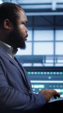 Dikey video Afrikalı Amerikalı adam yüksek teknolojili bir tesiste yürüyor, ağ sorunlarını çözmek için dizüstü bilgisayar kullanıyor ve yavaş veri transfer oranlarına yol açıyor. Veri merkezi çalışanı süper bilgisayarları etkileyen hataları düzeltiyor