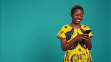 Modern genç bir kadın telefonuyla internet sitelerine girip mavi arka planda duruyor. Neşeli tatmin olmuş kız, stüdyoda akıllı telefon kullanarak sosyal medyada mesajlarını kontrol ediyor. Kamera B.