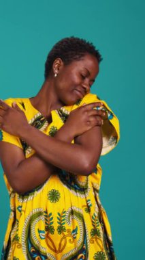 Dikey video özgüvenli genç kadın Afrikalı kökenini temsil ediyor. Stüdyoda çiçekli bir kabile elbisesi giyerek. Tatlı doğal kız kökeniyle gurur duyarak kendini kucaklıyor.