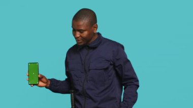 Gülümseyen güvenlik görevlisi koruma işinde sunum yapıyor akıllı telefon, stüdyo arkaplanı. Stüdyonun arka planında izole edilmiş yeşil ekranlı telefon gösteren neşeli koruma portresi.
