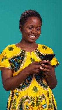 Dikey Video Modern kadın telefonuyla internet sitelerine girip mavi arka planda duruyor. Neşeli tatmin olmuş kız, stüdyoda akıllı telefon kullanarak sosyal medyada mesajlarını kontrol ediyor. Kamera.