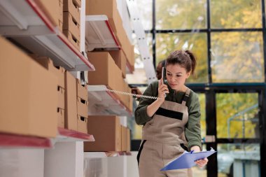 Beyaz bir kadın sabit telefon başında uzaktan kumandayla konuşuyor, depoda ürün kalitesini tartışıyor. Müşteri siparişlerinde çalışan çalışanlar, karton kutuları sevkiyat için hazırlıyorlar.