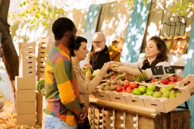 Kadın tüccar, çiftçi pazarında taze biyo-meyve ve sebze satarken müşterilere numune sunuyor. Genç çok ırklı aile mevsimlik organik ürünler deniyorlar, yemek festivalini ziyaret ediyorlar.