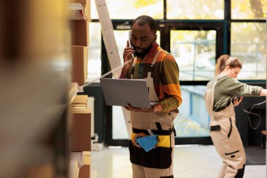 Depo çalışanı sabit telefon hattında uzaktan yöneticiyle konuşuyor, dizüstü bilgisayardaki müşterilerin siparişlerine bakıyor. Afro-amerikan yönetici ambalajları depoya teslim etmek için hazırlıyor.