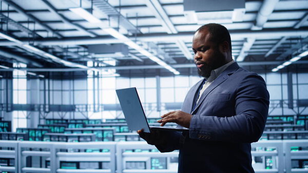 Programmer in data center typing on laptop, running error checking utilities. IT professional in server room looking for network issues affecting rigs performance using digital device, camera B