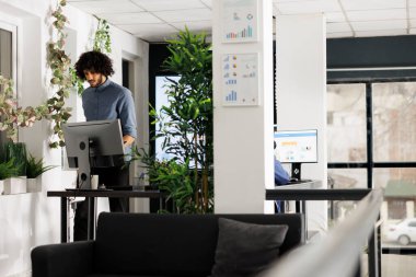 Arap şirket çalışanı yeni açılan uzay ofisinde yürüyor. Genç bir şirket çalışanı çalışma masasının yanında duruyor ve modern yeşil çalışma alanında bilgisayarı var.