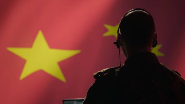 Çin ordusunun harekat subayı laptoptaki izleme iletimine bakıyor. Kulaklık kullanarak askerlere bilgi aktarıyor. Dalgalanan Çin bayrağı arka planında izole edilmiş.