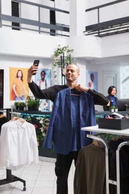 Gülümseyen Arap adam askıda ceket tutuyor ve elbise mağazasından alışveriş yaparken akıllı telefondan selfie çekiyor. Genç vlogger butikte cep telefonuyla marka tanıtım videosu kaydediyor