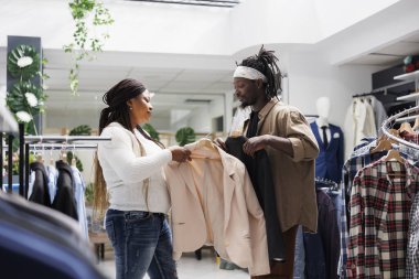 Kadın alışveriş merkezinde kıyafet seçerken adam askıda kıyafet tutuyor. Moda mağazasında kıyafet ararken gündelik modaya uygun ceketlere bakan Afrikalı Amerikalı arkadaşlar.