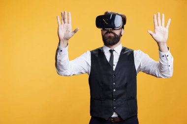 Modern garson sanal gerçekliği kullanıyor, sarı arka planda 3D VR kulaklığın keyfini çıkarıyor. Genç adam kamera önünde etkileşimli görüş simülasyonuyla eğleniyor, yenilikçi gözlükler.