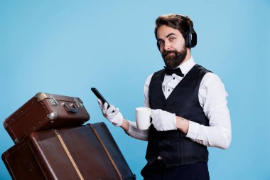 Hotel guard text and drinks coffee, checks emails on his mobile phone while he listens to tunes. Bellman employee navigating internet pages and sipping refreshing drink with headset on. clipart