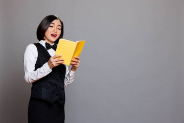 Asian waitress in uniform reading novel during break at restaurant. Catering service young attractive woman employee holding book and enjoying literature with surprised face expression clipart