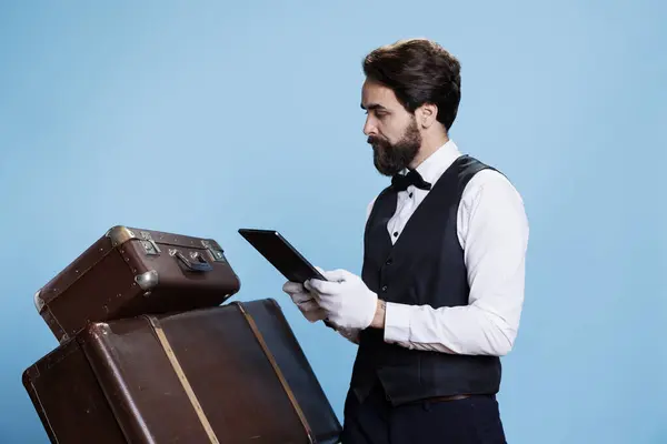 stock image Bellhop verifying luggage for guests and checking tablet with online bookings, standing against blue background. Classy hotel porter with suit and tie reviews reservations on device, tourism concept.