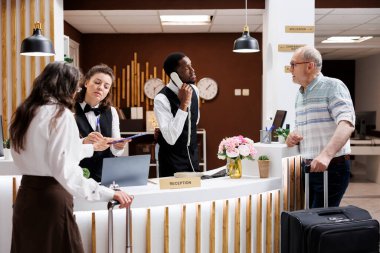 Otel resepsiyonistleri yaşlı yolculara kayıt masasında yardım ediyor. Müteşekkir kıdemli kadın evrakları gülümseyerek imzalarken çalışanlar süreci kolaylaştırıyor. Olağanüstü müşteri hizmetleri.