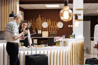 Son sınıf erkek turist otel resepsiyonuna giriş yapıyor, eğlenceli bir tatil için hazır. Dost canlısı bayan kapıcı yaşlı adam rezervasyon formlarını doldururken ona yardımcı oluyor..