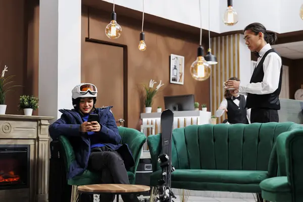 stock image In lounge area, woman in winter jacket is using her smartphone as young server gives her a coffee. Female tourist using a mobile device to plan activities during her skiing trip at a fancy winter