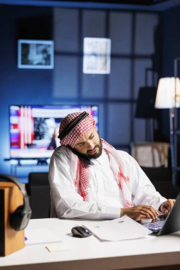 Arab entrepreneur in traditional attire works in office browsing on laptop and speaking on cell phone. Muslim man exemplifies professionalism and communication skills by utilizing modern technology. clipart