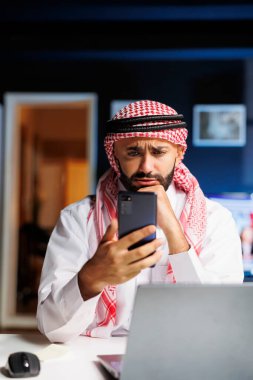 Geleneksel kıyafetler giymiş Müslüman bir adam ofis masasında video görüşmesi için mobil cihaz kullanıyor. Çevrimiçi araştırma ve görevler için akıllı telefonunu kavrıyor, kendinden emin ve işiyle meşgul görünüyor..