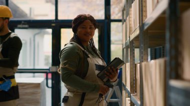 Afrika kökenli Amerikalı lojistik şefi portresi, eşyaları tarıyor ve çevrimiçi ekommerce web sitesi için fiyatları güncellerken, arka plandaki stajyer karton kutuların taşınması için hazır bekliyor.