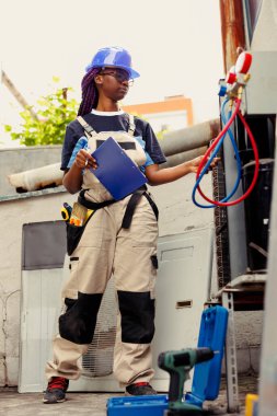 Adept repairman commissioned to optimize HVAC system efficacy, ensuring it operates at maximum capability. Proficient worker doing condenser unit investigation, imputing data on laptop clipart