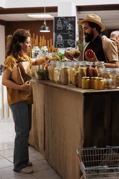 stock image Client shopping for organic pantry staples in zero waste store using biodegradable paper bag, being assisted with information by trader. Vendor showing woman products in reusable jars