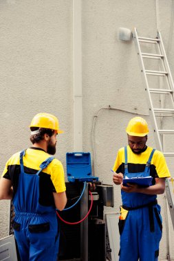 Uzman işçiler hvac sistemindeki sıvı ve gazların basıncını okumak için manifold göstergeleri kullanırken, kalifiye elektrikçiler açık hava kondansatör denetimi ve güvenlik denetimi sonuçlarını panoya yazıyorlar.