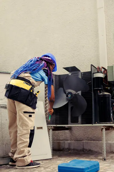 stock image African american skilled mechanic commissioned by home owner for annual air conditioner routine maintenance, using soft dusting brush to clean dusty condenser coils and filters, outdoor