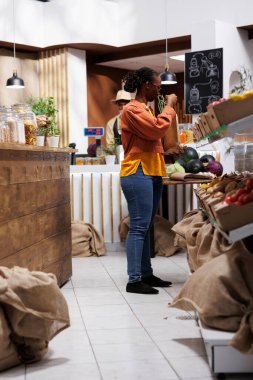 Siyahi kadın, çevre dostu, sürdürülebilir ürünler için modern bir marketten alışveriş yapıyor. Yerel sebzeleri buluyor ve yeniden kullanılabilir poşetler alıyor. Mağaza çevre dostu organik gıdalar sunuyor..