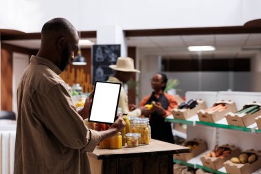 Afrikalı Amerikalı erkek müşteri dikey olarak izole bir kromakey model şablonuyla dijital tableti kavrıyor. Mağazada yeniden kullanılabilir ambalaj ve sürdürülebilir ürünler ahşap raflarda bulunuyor.