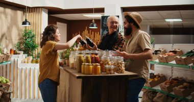 Senior vendor assisting clients in eco friendly shop with ethical sourced chemicals free food items. Couple in local store chatting with trader about pantry staples bulk products, zoom out clipart