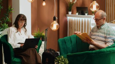 Üst düzey çift bekleme salonunda bekliyor, kontrol sürecinden geçmeden önce farklı rahatlatıcı aktivitelerle zaman geçiriyorlar. Kadın dizüstü bilgisayarda internette geziniyor ve erkek otelde kitap okuyor..