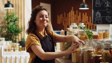 Gülümseyen sıfır atık süpermarket sahibinin portresi iklim etkisini azaltmak için ayrıştırılabilir cam kaplar kullanıyor. Yerel bakkaldaki neşeli kadın, malları düzenliyor.