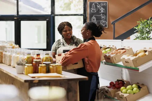 Stock image Black woman wearing an apron is carrying boxes of fresh pesticide free produce from local farmer in eco friendly store. Female supplier delivers crates of farm grown products to bio food shop owner.