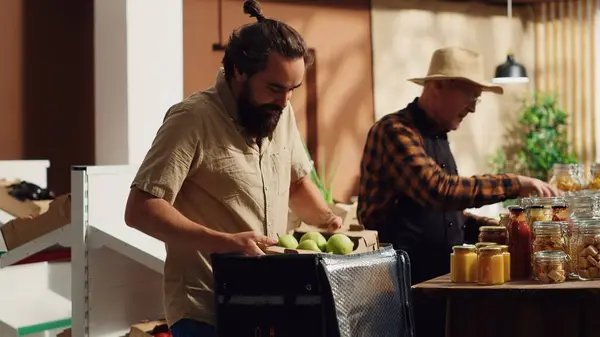 stock image Deliveryman providing organic zero waste supermarket food orders to customers, helped by aged merchant to fill thermic backpack. Man bringing local shop chemicals free groceries to customers