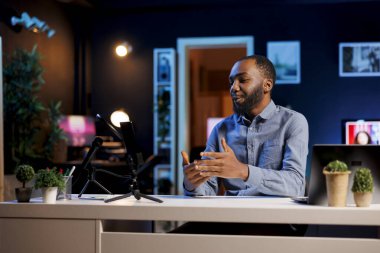 Hevesli BIPOC internet şovu sunucusu cep telefonu tutucusuna bağlı akıllı telefondaki kayıt düğmesine basıyor, mikrofonu bugünün video konusunu abonelerle konuşmak için kullanıyor