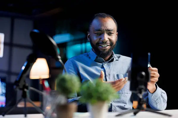 stock image BIPOC content creator presenting phone from brand sponsoring video, urging viewers to buy it. Viral media star does influencer marketing by advertising cellphone to audience