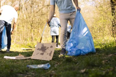 Genç gönüllü, ormanı çöp ve plastikten temizlemek için uzun pençeli bir aletle çöp topluyor, maşayla çöp topluyor. Eylemci, yasadışı boşaltmayla mücadele etmek için gönüllü iş yapıyor..