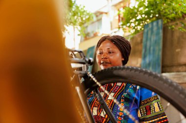 Genç hevesli zenci kadın dışarıda modern bisikletin bakımını ve bakımını yapıyor. Aktif Afro-Amerikan kadın bisikletçi dışarıda motorsikleti kontrol ederken görüldü..