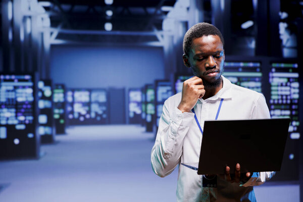 Electrician inspecting blade servers delivering online web content to customers. African american wireman using laptop to take note of critical systems, looking for damaging errors