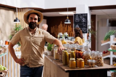 Çevre dostu sıfır atık mağazasında sebze arayan gülümseyen bir adamın portresi. Hipster müşterisi market alışverişi yapıyor, çevre dostu mahalle mağazasından vejetaryen yemekleri alıyor.