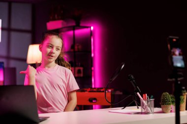 Hoş kız içerik yaratıyor, cep telefonunu tripod dengeleyicisinde kullanarak vlog kanalı girişini çekiyor. Gen Z eğlendiricisi, bugün video konusunu sunan, cep telefonuyla kayıt yapan çocuk seyirciyi selamlıyor