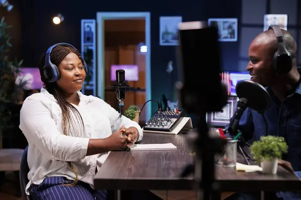 stock image Influential African American host engages with guest, using quality podcast equipment in their stylish home studio. Black couple utilizing smartphone on tripod to record their online talk show.