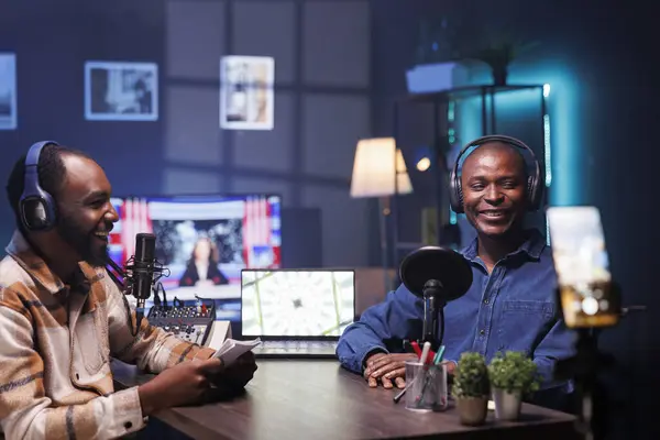 stock image Cheerful vloggers streaming podcast using smartphone on tripod during live debate on vlog channel. Excited men meeting in studio to film show with mobile phone and talk global events.