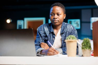 Odaklanmış Afro-Amerikan kadın evde çalışıyor, internette geziniyor, çevrimiçi çalışıyor ve modern oturma odasında birden fazla iş yapıyor. Dişi blog yazarı dizüstü bilgisayara bakarken elinde kalem tutuyor.