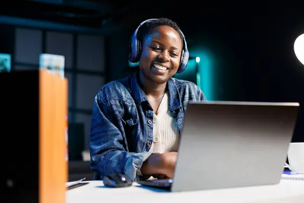 stock image Cheerful black woman uses wireless headphones while browsing the internet, and attending virtual meetings on her laptop. African american lady multitasks, streaming movies and studying online.