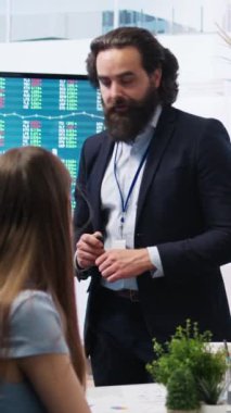Dikey video finans danışmanı, büyük ekran kullanarak müşterilere pazar analizleri sunarak, veriye dayalı karar verme sürecini vurguluyor. Borsa göstergeleri hakkında istatistiksel çizelgeler gösteren broker