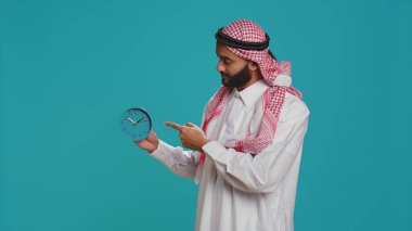 Islamic adult looks at wall clock to check time on camera, using timepiece to see hour and minutes. Young person in arabic national costume checking local timezone, blue watch decoration. clipart